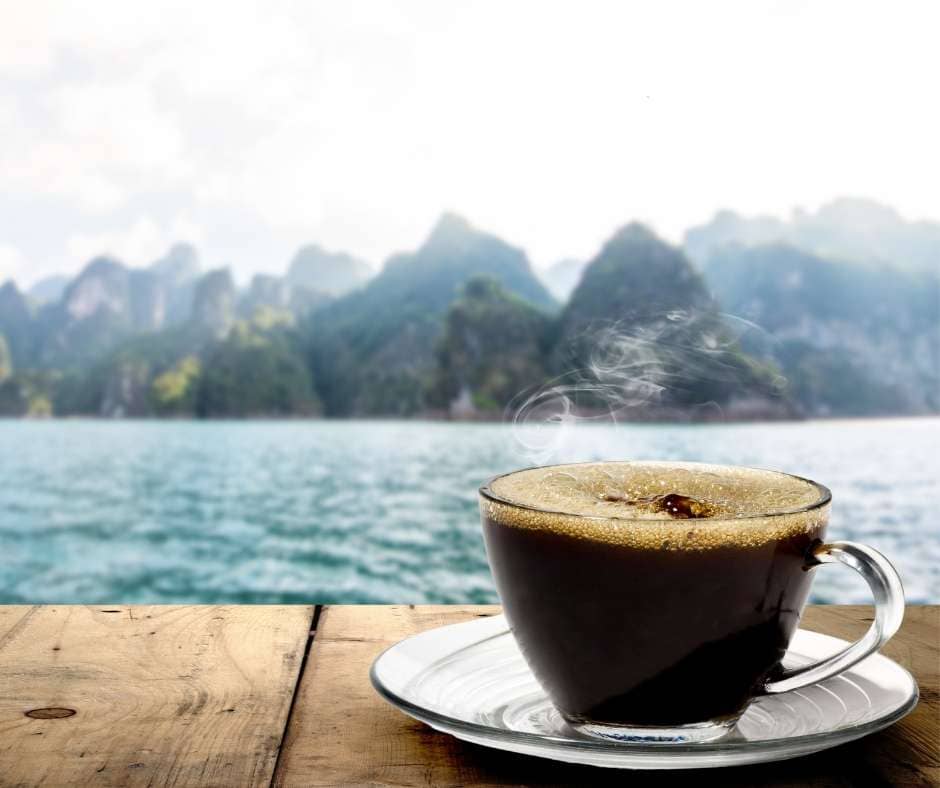 Steaming cup of freshly roasted American coffee in a glass cup on wooden table by the water with mountains