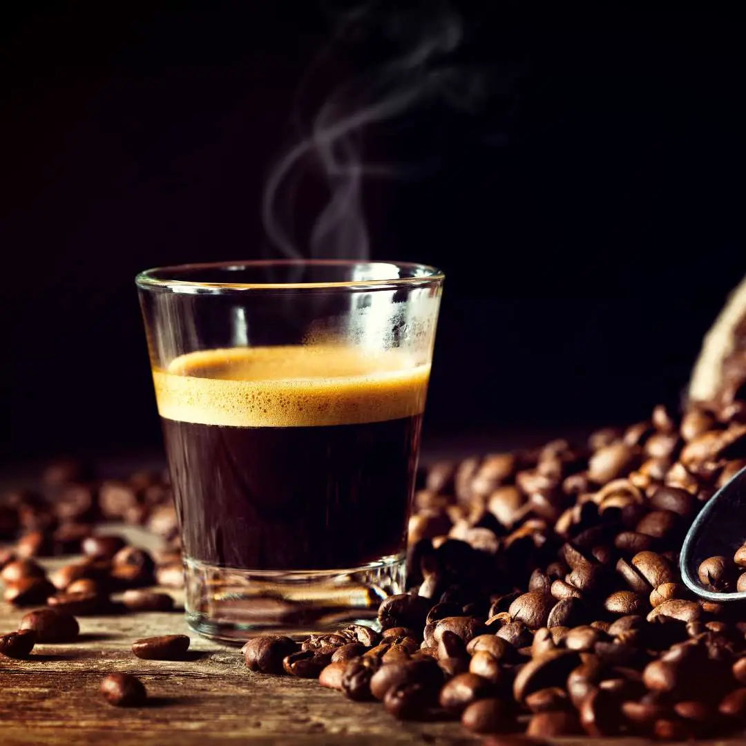 Glass espresso shot with crema and steam on wooden table surrounded by beans Caffeeegy Co locally roasted coffee