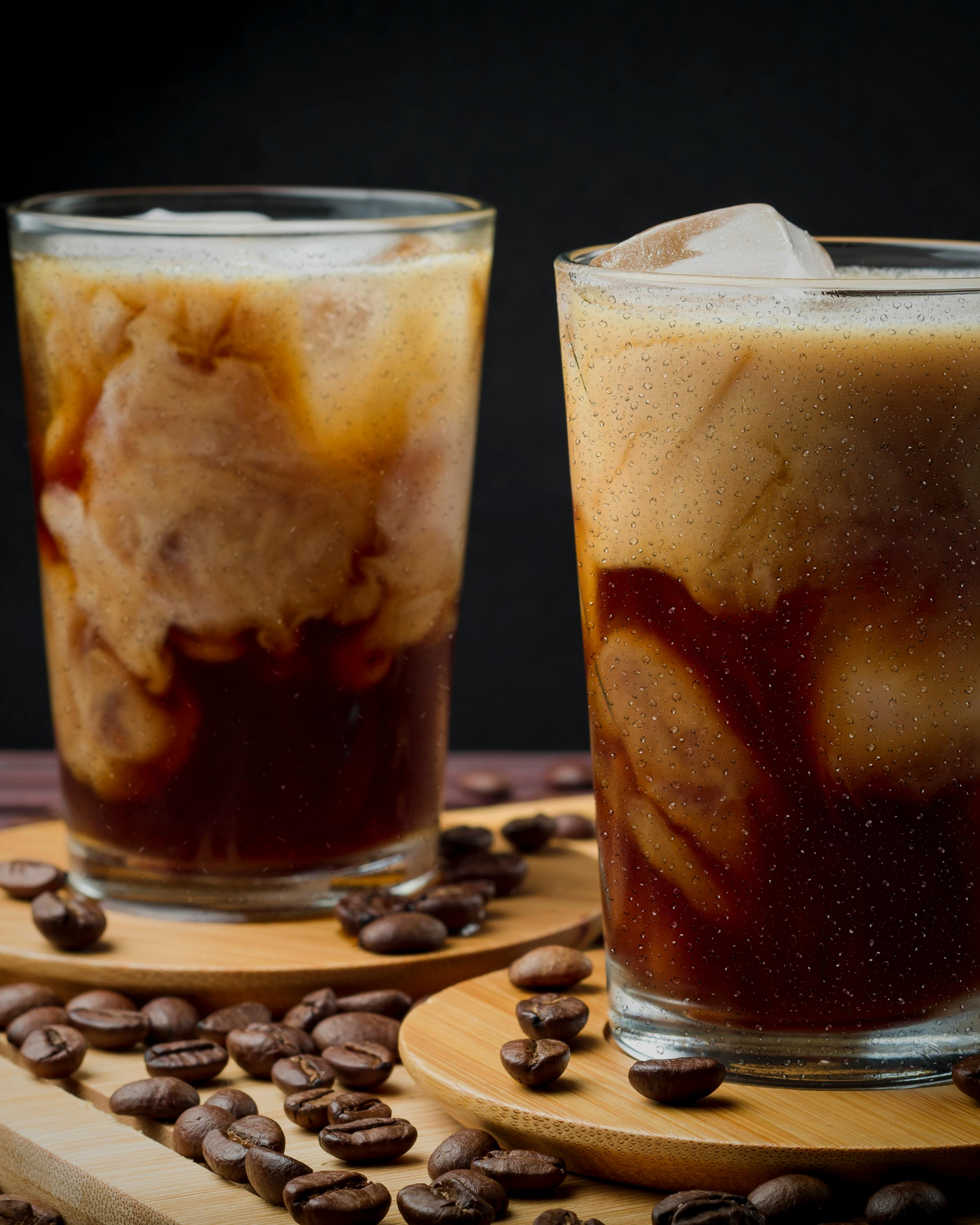Two glasses of iced coffee made with premium coffee powder surrounded by coffee beans on wooden coasters